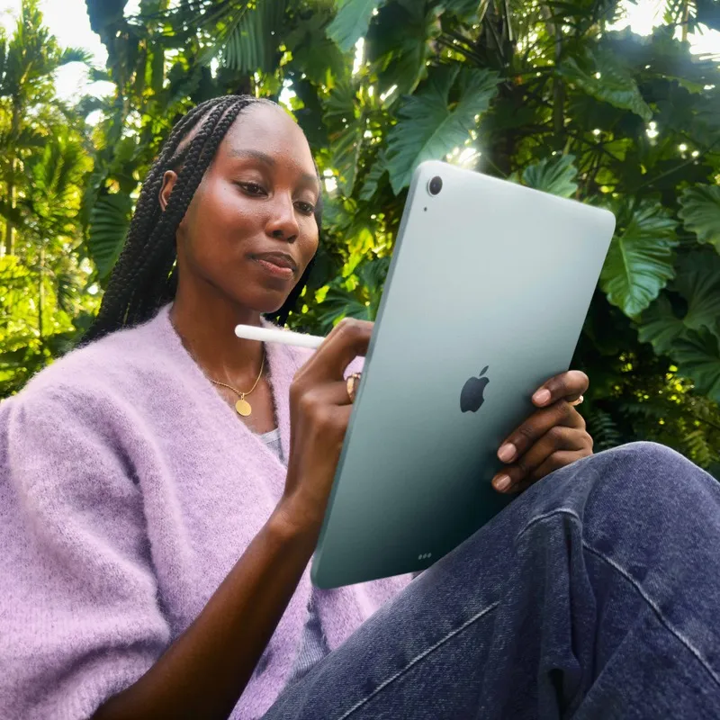 Woman using a blue Apple iPad Air 13-inch M4 512GB tablet with a stylus in a lush outdoor setting at YourShoppy.com.