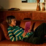A person relaxes on a brown leather sofa holding a pink Apple iPad (A16) Wi-Fi 256GB tablet at YourShoppy.com in Bangalore.